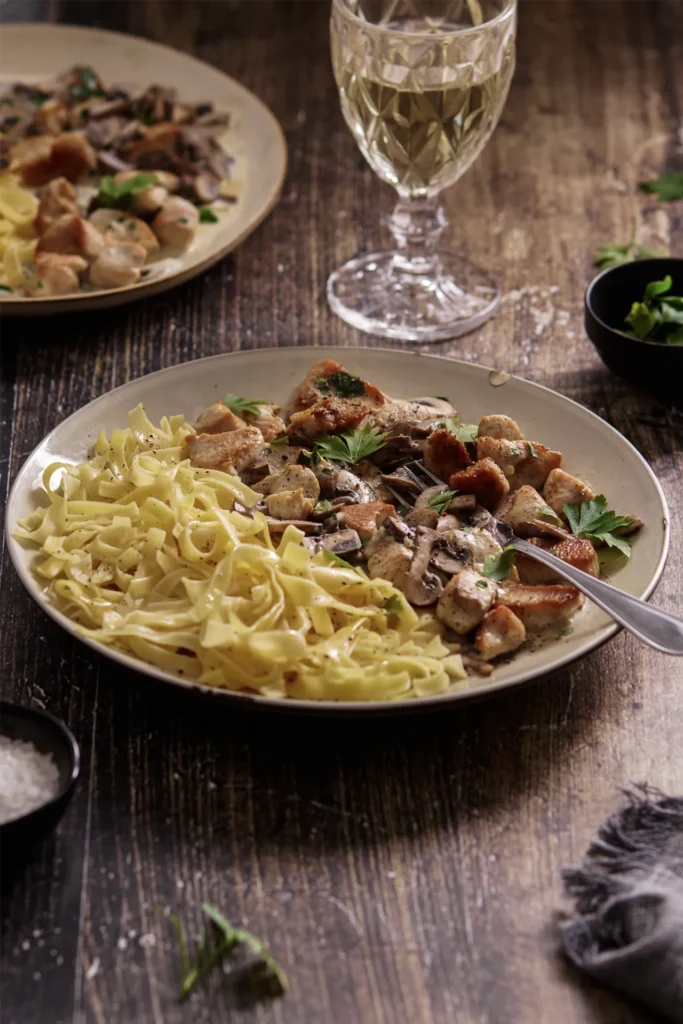 Puten-Geschnetzeltes mit Champignons in cremiger Sauce, serviert mit Bandnudeln auf einem Teller, garniert mit frischer Petersilie.