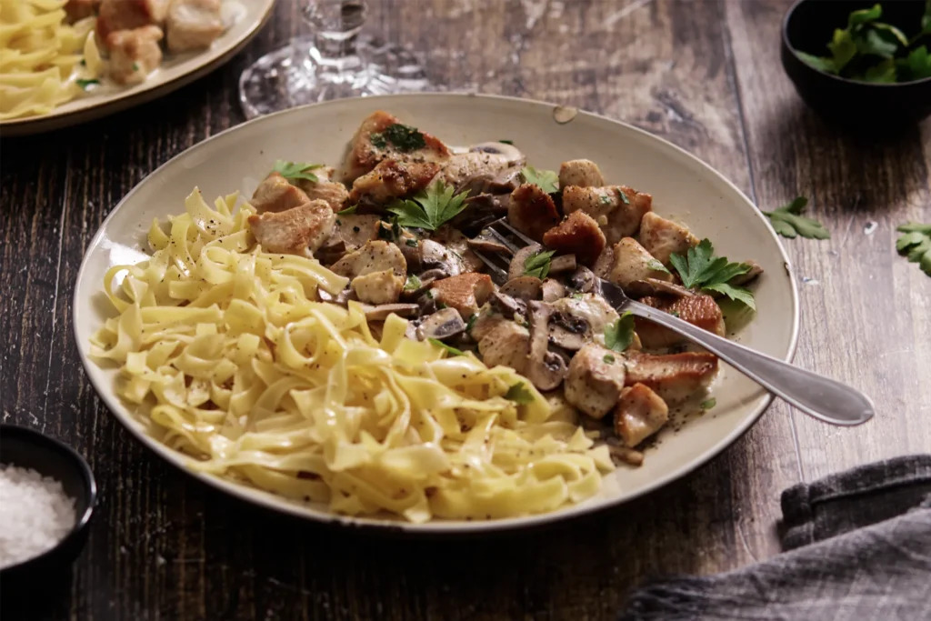 Puten-Geschnetzeltes mit Champignons in cremiger Sauce, serviert mit Bandnudeln auf einem Teller, garniert mit frischer Petersilie.